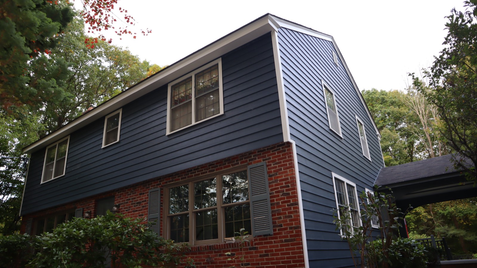 Colonial corner clad in navy siding over a brick base with a wooded backdrop